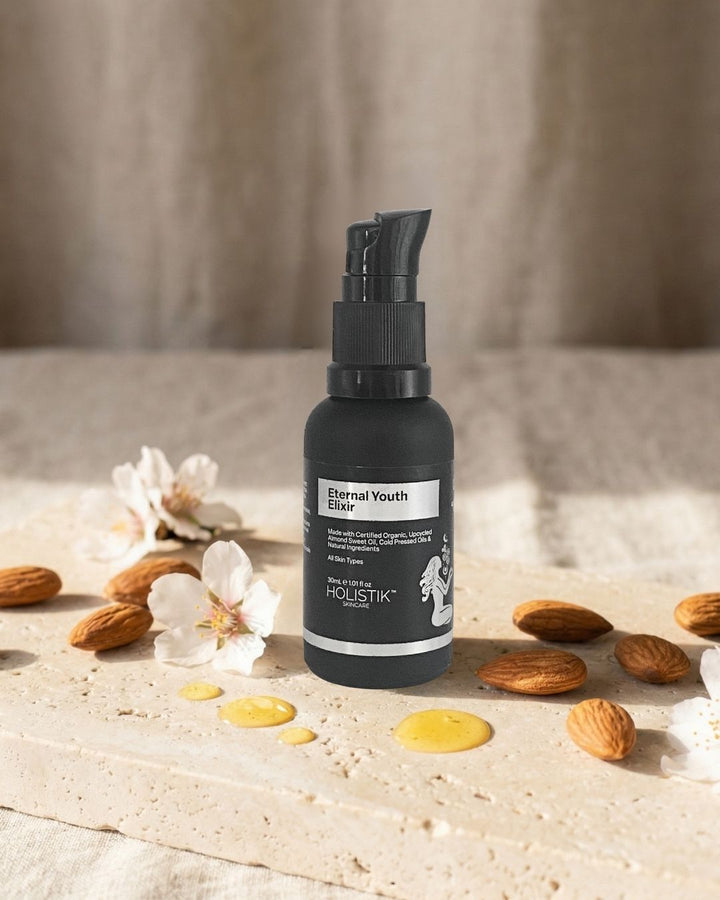 Black bottle of 'Eternal Youth Elixir' with almonds and flowers on a beige surface