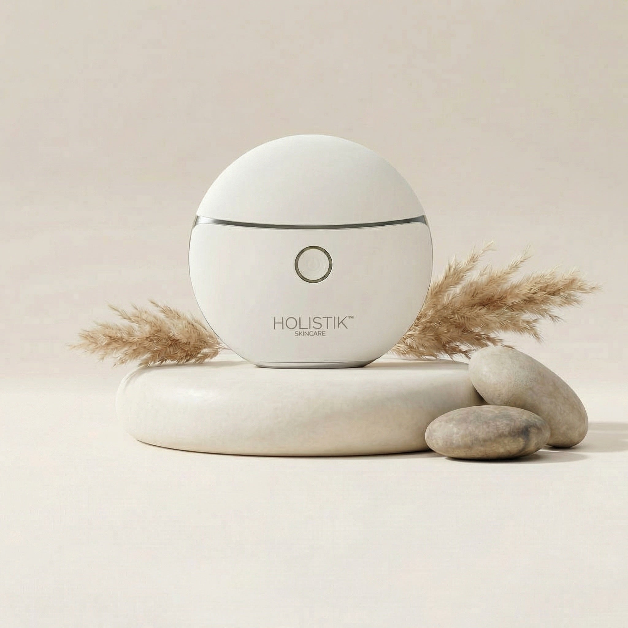 White branded LED Gua Sha Face & Body massage device from Holistik skincareon a beige background with stones and dried plants.