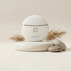 White branded LED Gua Sha Face & Body massage device from Holistik skincareon a beige background with stones and dried plants.