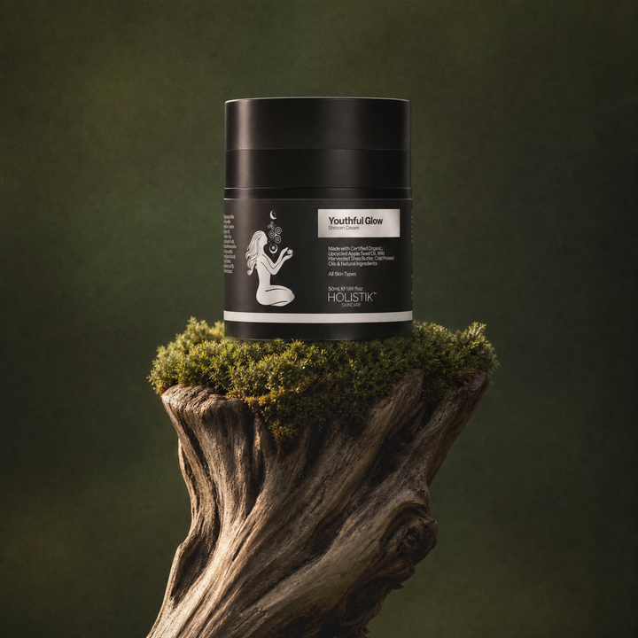 A black cosmetic jar labeled 'Youthful Glow Shroom Cream' and 'Holistik Skincare' sits on a mossy wooden stump against a dark olive-green background.